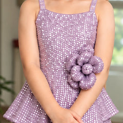 A child wearing a lavender sequined sleeveless dress with a large matching fabric flower embellishment on the front, standing indoors with hands folded.