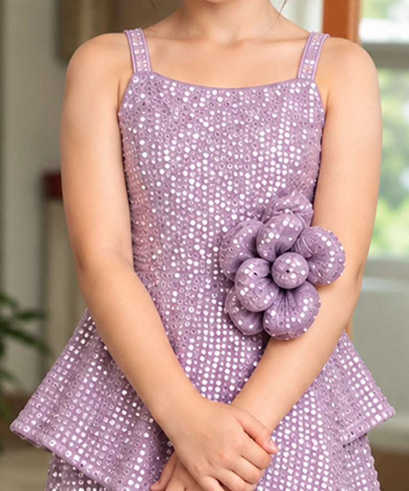 A child wearing a lavender sequined sleeveless dress with a large matching fabric flower embellishment on the front, standing indoors with hands folded.