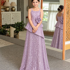 A young girl wearing a lavender sequined formal gown with a ruffled peplum and a matching flower applique on the bodice, standing in an indoor setting with a mirror showing her reflection.