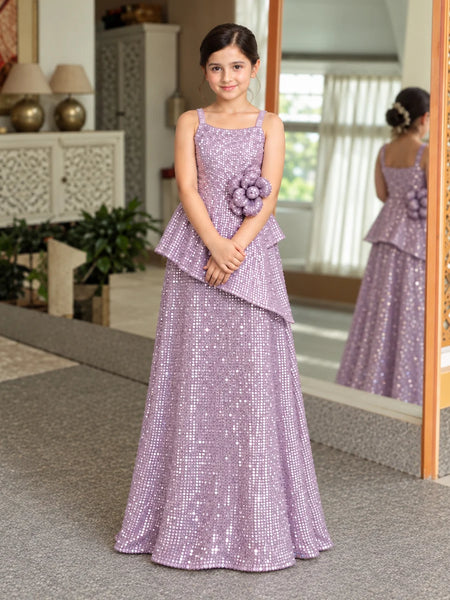 A young girl wearing a lavender sequined formal gown with a ruffled peplum and a matching flower applique on the bodice, standing in an indoor setting with a mirror showing her reflection.