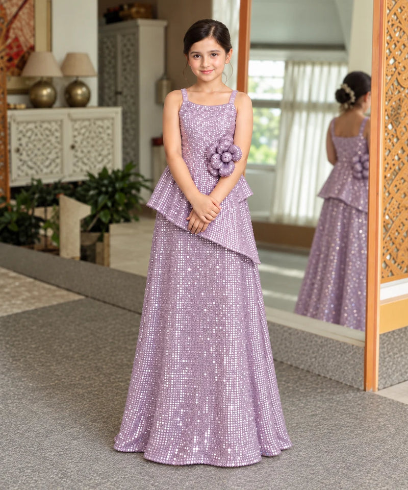 A young girl wearing a lavender sequined formal gown with a ruffled peplum and a matching flower applique on the bodice, standing in an indoor setting with a mirror showing her reflection.
