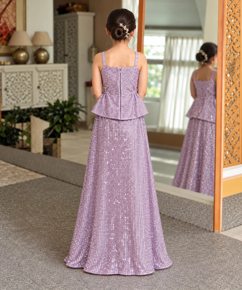 A young girl viewed from behind wearing a lavender purple sequined formal gown with a peplum waist, standing in a decorated indoor hallway with a mirror reflecting her profile.