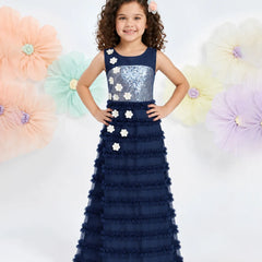 A young girl with curly brown hair wearing a navy blue formal gown with a sequined bodice and ruffled skirt, standing against a white background with large pastel paper flowers.