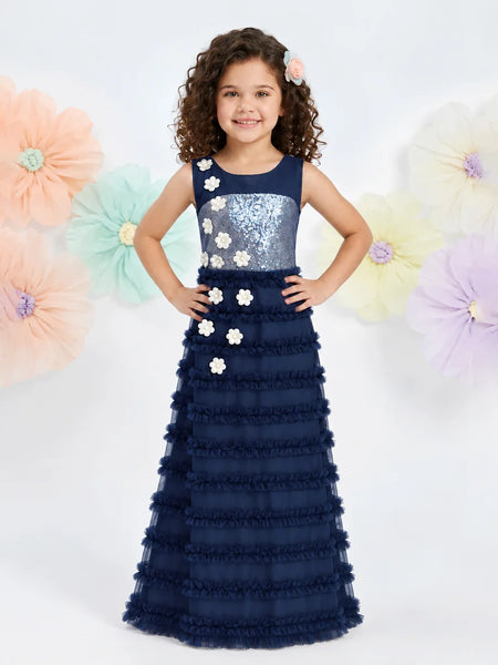 A young girl with curly brown hair wearing a navy blue formal gown with a sequined bodice and ruffled skirt, standing against a white background with large pastel paper flowers.