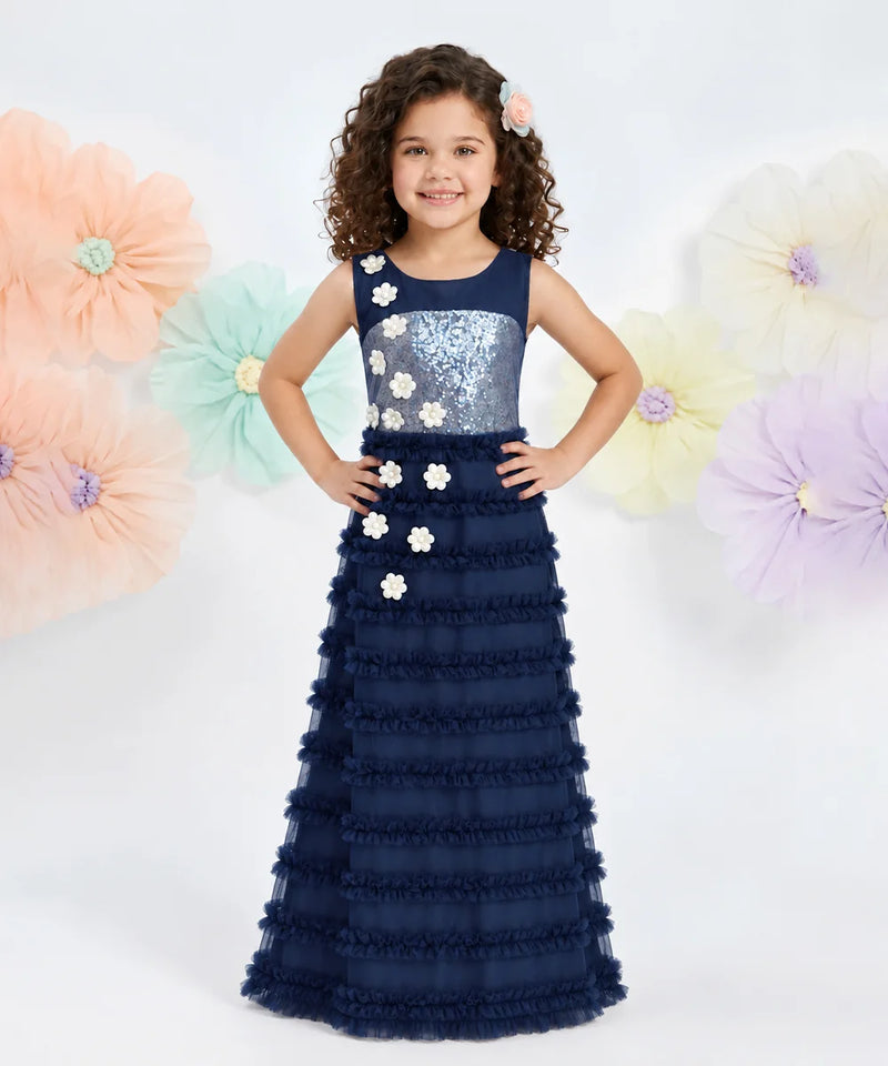 A young girl with curly brown hair wearing a navy blue formal gown with a sequined bodice and ruffled skirt, standing against a white background with large pastel paper flowers.