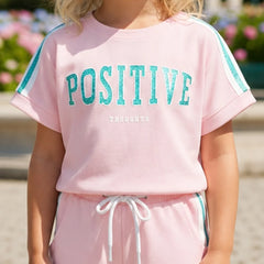 A child wearing a light pink short-sleeve sweatshirt with 'POSITIVE THOUGHTS' printed in teal across the chest, paired with matching pink sweatpants with a white drawstring and teal side stripes.