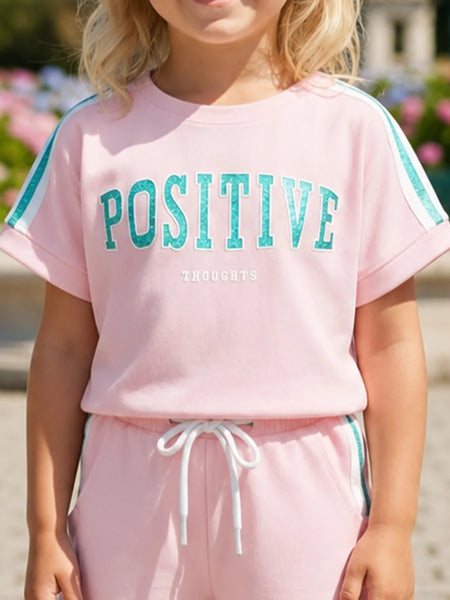 A child wearing a light pink short-sleeve sweatshirt with 'POSITIVE THOUGHTS' printed in teal across the chest, paired with matching pink sweatpants with a white drawstring and teal side stripes.
