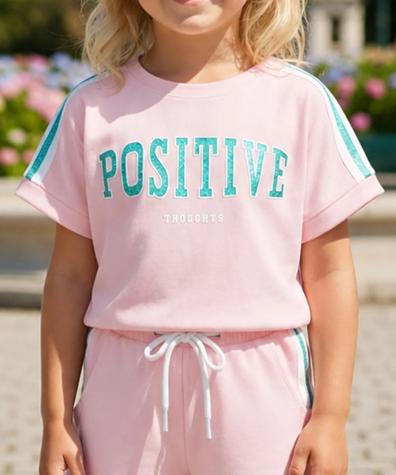 A child wearing a light pink short-sleeve sweatshirt with 'POSITIVE THOUGHTS' printed in teal across the chest, paired with matching pink sweatpants with a white drawstring and teal side stripes.