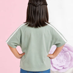 A child shown from the back wearing a sage green short-sleeve t-shirt with white stripes on the shoulders, paired with blue jeans, set against a split pink and white background with a large purple flower prop.