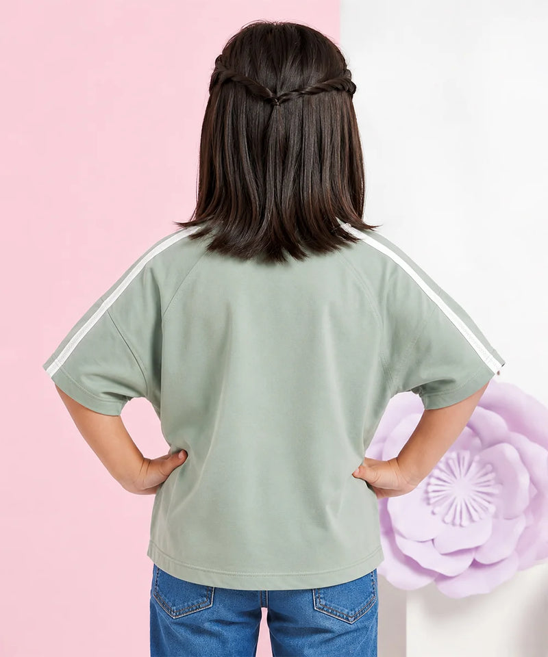 A child shown from the back wearing a sage green short-sleeve t-shirt with white stripes on the shoulders, paired with blue jeans, set against a split pink and white background with a large purple flower prop.