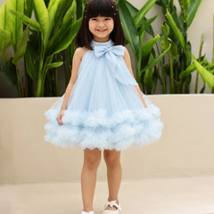 A young girl with dark hair wearing a light blue sleeveless tulle dress with a large bow at the neckline and a fluffy tiered skirt, standing outdoors in front of green plants and a beige wall.