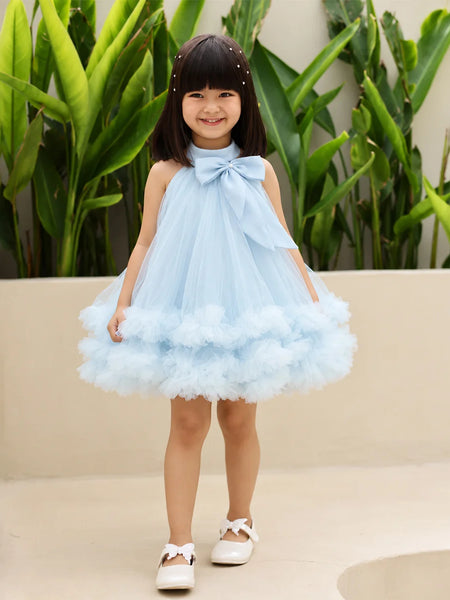 A young girl with dark hair wearing a light blue sleeveless tulle dress with a large bow at the neckline and a fluffy tiered skirt, standing outdoors in front of green plants and a beige wall.