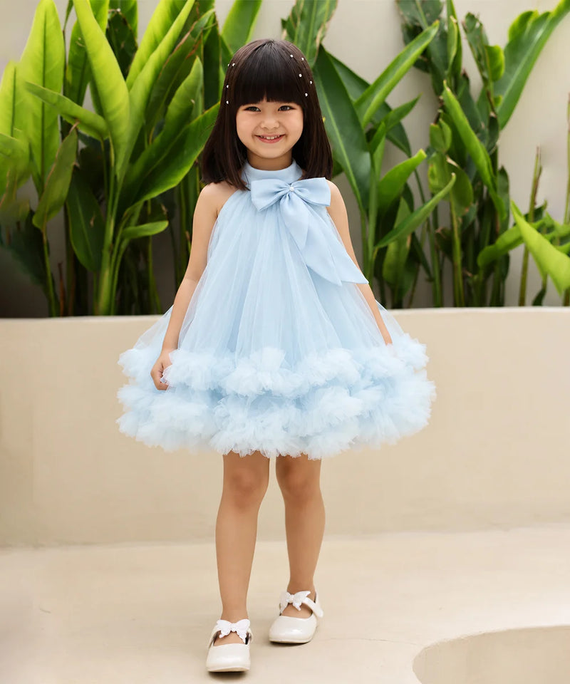 A young girl with dark hair wearing a light blue sleeveless tulle dress with a large bow at the neckline and a fluffy tiered skirt, standing outdoors in front of green plants and a beige wall.