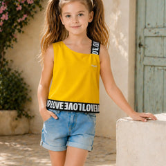 A young girl with pigtails stands outdoors wearing a bright yellow sleeveless top with black lettering on the straps and hem, paired with light blue denim shorts.
