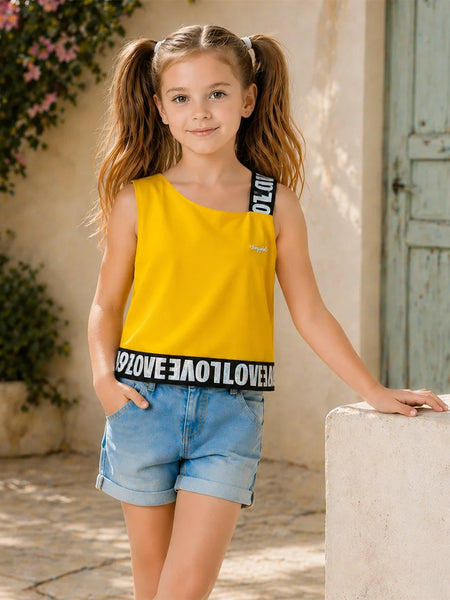 A young girl with pigtails stands outdoors wearing a bright yellow sleeveless top with black lettering on the straps and hem, paired with light blue denim shorts.