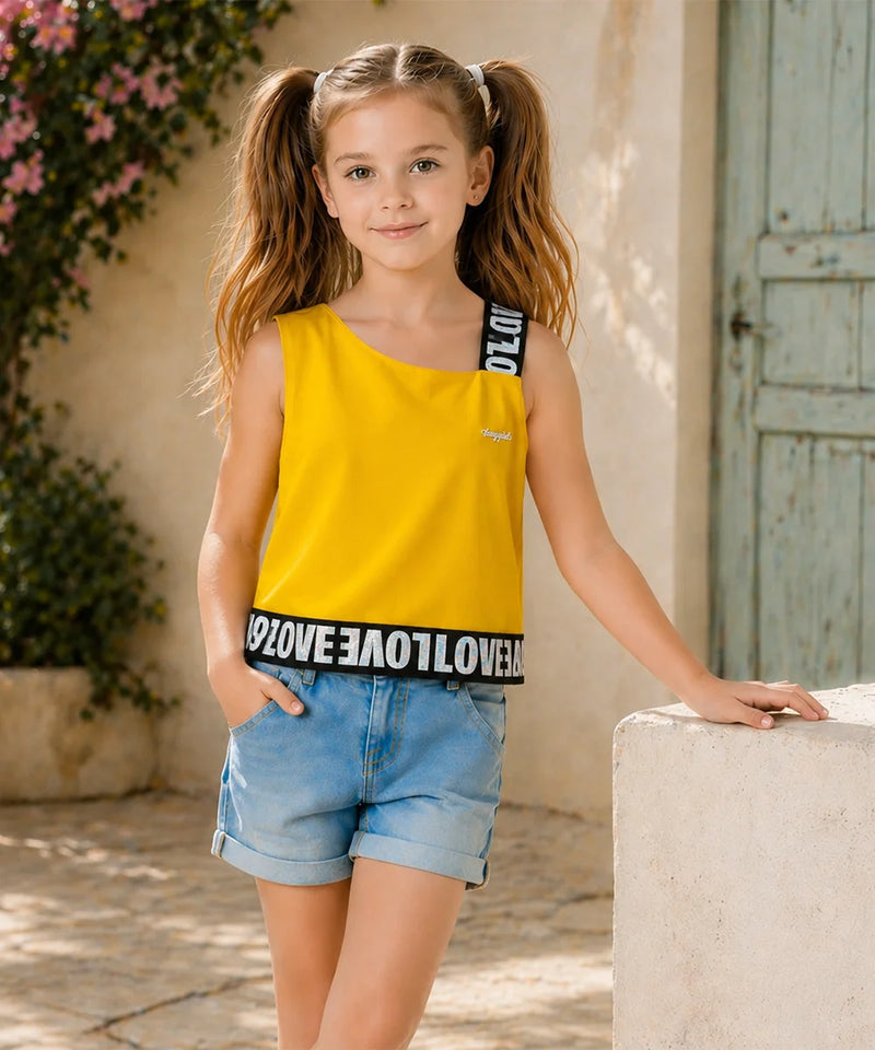 A young girl with pigtails stands outdoors wearing a bright yellow sleeveless top with black lettering on the straps and hem, paired with light blue denim shorts.