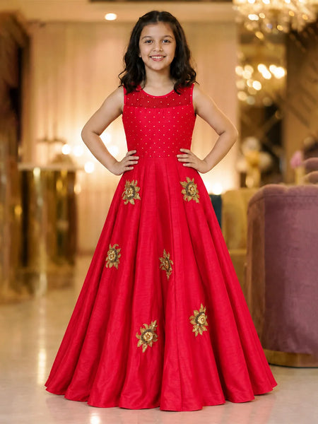 A young girl stands in a grand, warmly lit indoor hallway wearing a bright red sleeveless evening gown with gold floral embroidery and a subtle polka-dot pattern on the bodice.