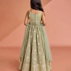 A young girl viewed from behind wearing a sage green traditional Indian gown with gold embroidery and a matching sheer dupatta, set against a warm peach studio backdrop.