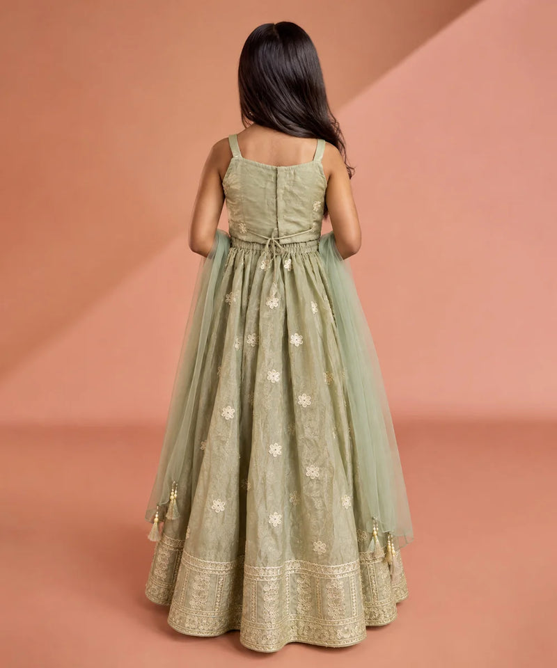 A young girl viewed from behind wearing a sage green traditional Indian gown with gold embroidery and a matching sheer dupatta, set against a warm peach studio backdrop.