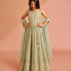 A young girl modeling a sage green anarkali gown with gold embroidery, a matching dupatta, and a gold tassel detail, set against a solid dusty pink background.