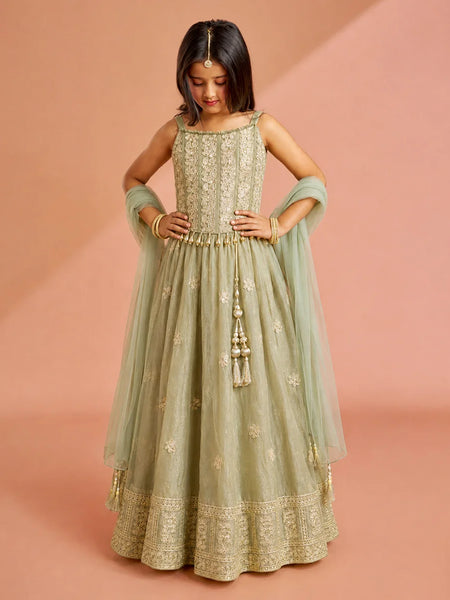 A young girl modeling a sage green anarkali gown with gold embroidery, a matching dupatta, and a gold tassel detail, set against a solid dusty pink background.