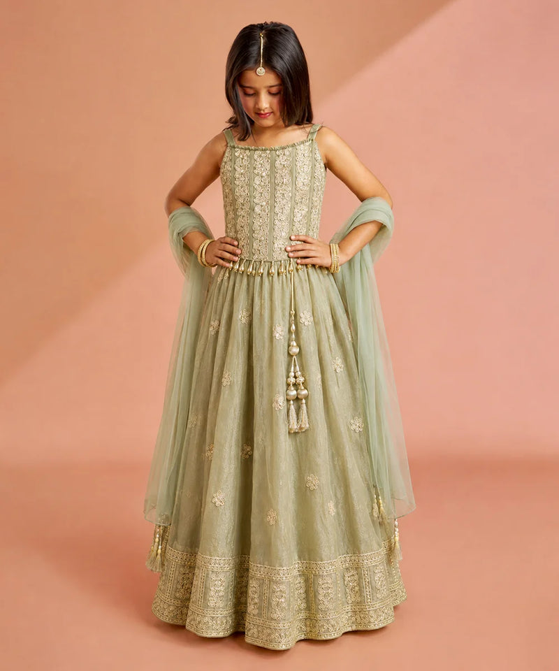 A young girl modeling a sage green anarkali gown with gold embroidery, a matching dupatta, and a gold tassel detail, set against a solid dusty pink background.