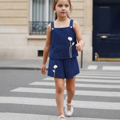 A young girl walking across a city crosswalk wearing a navy blue two-piece outfit with white embroidered flowers and white sneakers.