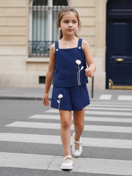 A young girl walking across a city crosswalk wearing a navy blue two-piece outfit with white embroidered flowers and white sneakers.