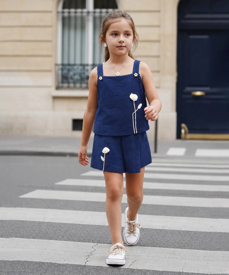 A young girl walking across a city crosswalk wearing a navy blue two-piece outfit with white embroidered flowers and white sneakers.