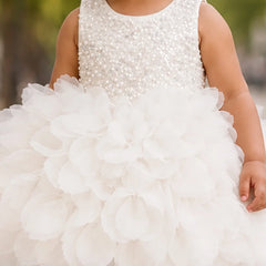 A child wearing a sleeveless white dress with a beaded embellished bodice and a large layered tulle flower skirt, photographed outdoors with a blurred green background.