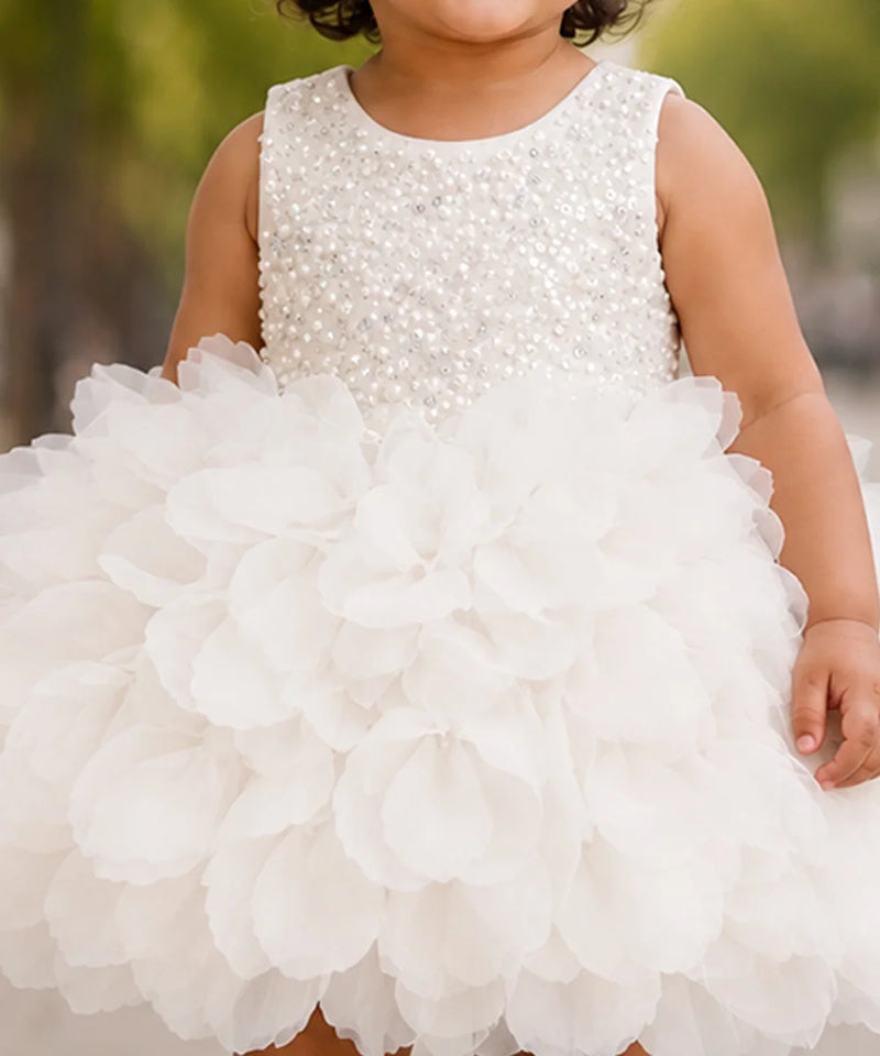 A child wearing a sleeveless white dress with a beaded embellished bodice and a large layered tulle flower skirt, photographed outdoors with a blurred green background.