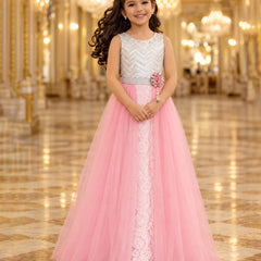 A young girl with long dark curly hair stands in a grand, ornate hall wearing a formal party dress. The dress features a silver and white chevron-patterned bodice, a silver waistband with a pink flower accent, and a voluminous pink tulle skirt with a white lace center panel.