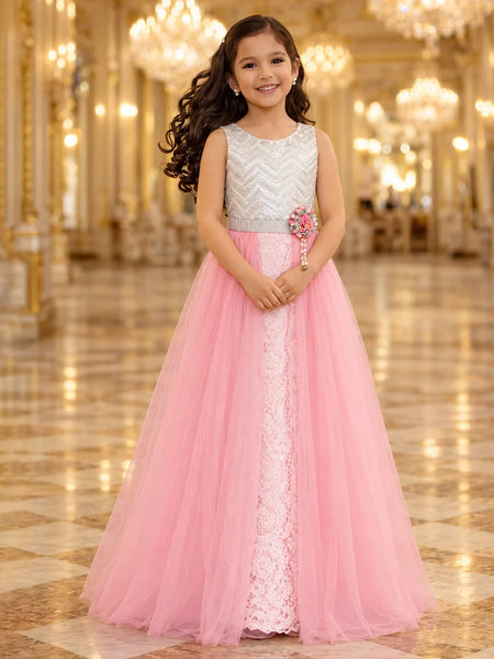 A young girl with long dark curly hair stands in a grand, ornate hall wearing a formal party dress. The dress features a silver and white chevron-patterned bodice, a silver waistband with a pink flower accent, and a voluminous pink tulle skirt with a white lace center panel.