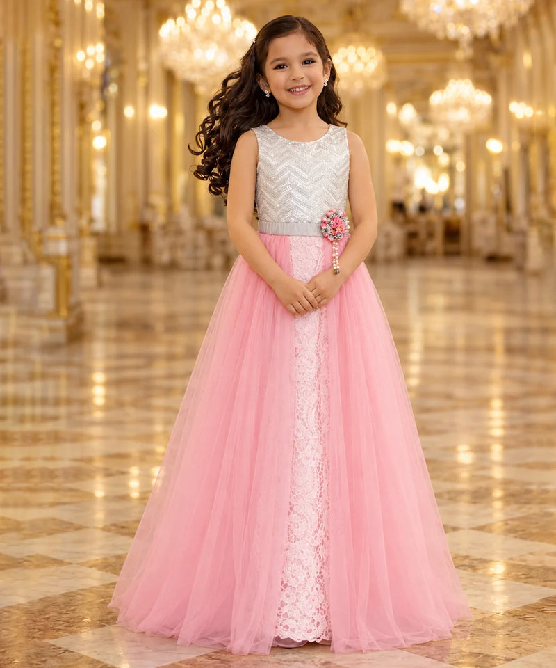 A young girl with long dark curly hair stands in a grand, ornate hall wearing a formal party dress. The dress features a silver and white chevron-patterned bodice, a silver waistband with a pink flower accent, and a voluminous pink tulle skirt with a white lace center panel.