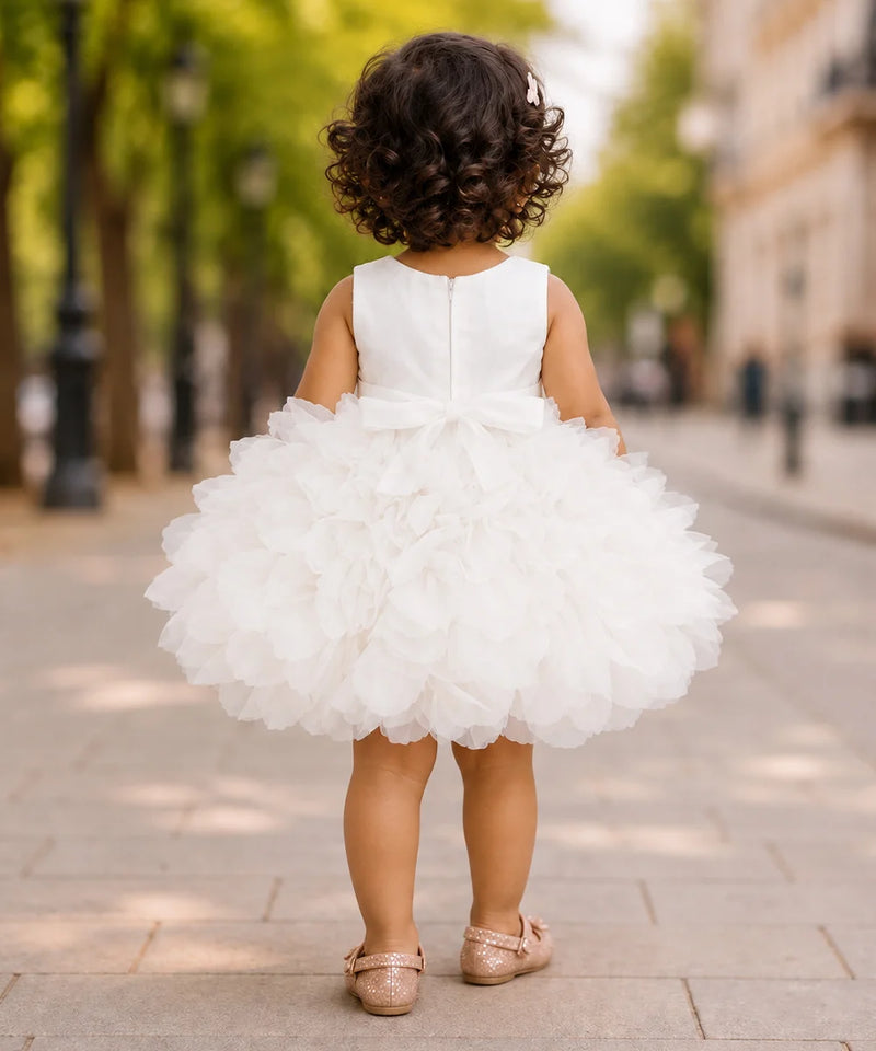A toddler girl viewed from behind wearing a sleeveless white tulle flower girl dress with a large bow at the back and a fluffy tiered skirt, standing on a paved street with blurred trees and buildings in the background.