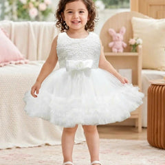 A toddler girl viewed from behind, wearing a sleeveless white tulle flower girl dress with a beaded bodice and a ruffled skirt, standing in a softly decorated room.