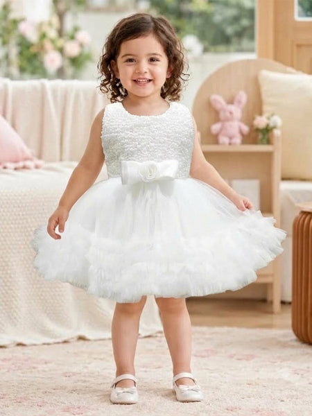 A toddler girl viewed from behind, wearing a sleeveless white tulle flower girl dress with a beaded bodice and a ruffled skirt, standing in a softly decorated room.