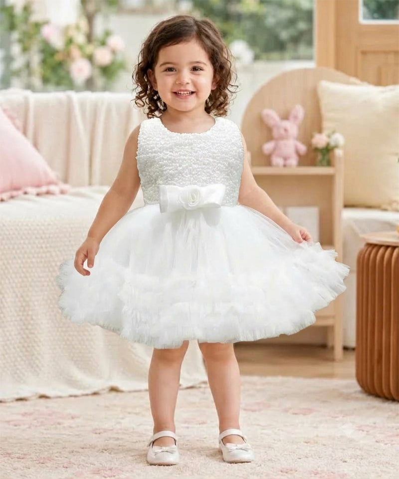 A toddler girl viewed from behind, wearing a sleeveless white tulle flower girl dress with a beaded bodice and a ruffled skirt, standing in a softly decorated room.