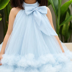 A young girl wearing a light blue sleeveless tulle dress with a large bow at the neckline and a ruffled tulle hem, photographed outdoors with green leaves in the background.