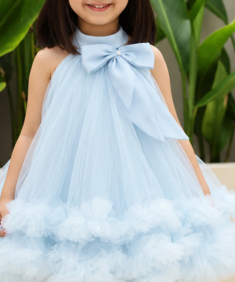 A young girl wearing a light blue sleeveless tulle dress with a large bow at the neckline and a ruffled tulle hem, photographed outdoors with green leaves in the background.