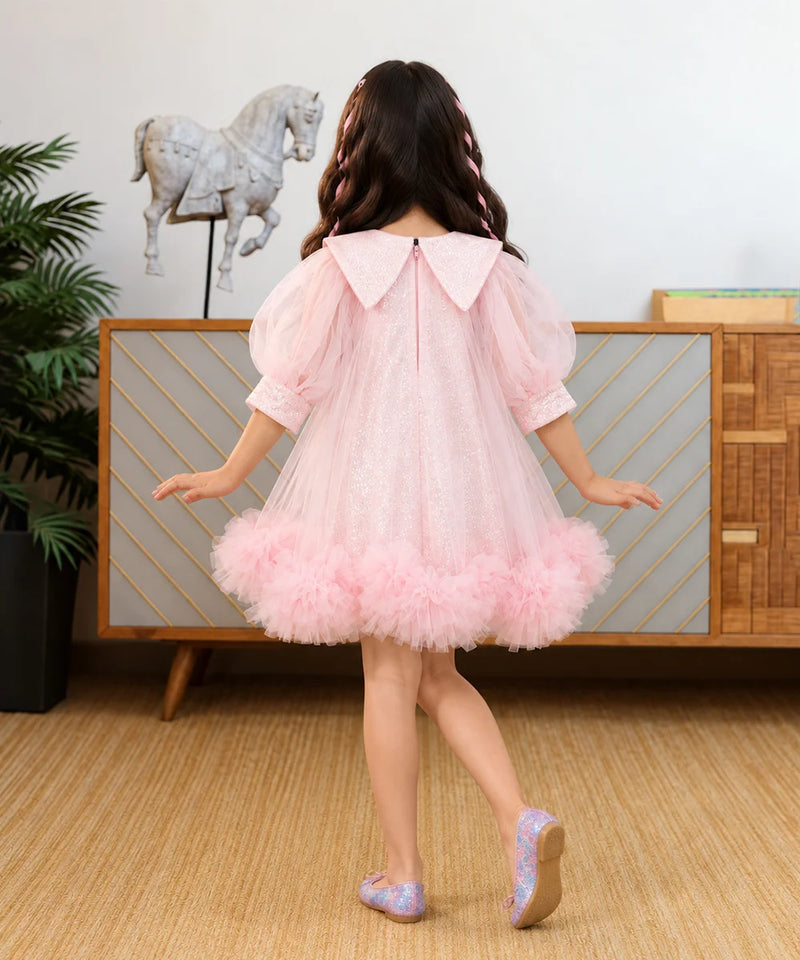 A young girl wearing a light pink sequined tulle dress with a large embellished Peter Pan collar, puff sleeves, and a ruffled tulle hem.