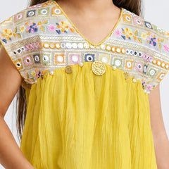 A close-up of a child wearing a bright yellow top with a heavily embroidered multicolor yoke. The embroidery features floral patterns and geometric borders in pink, blue, green, orange, and white, accented with small pearl-like beads and a central round gold-tone decorative button.