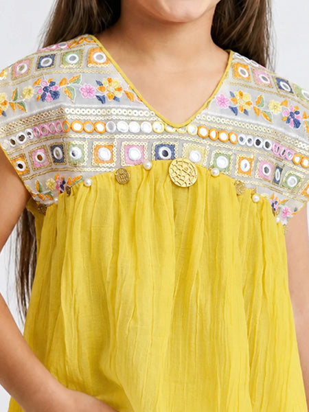 A close-up of a child wearing a bright yellow top with a heavily embroidered multicolor yoke. The embroidery features floral patterns and geometric borders in pink, blue, green, orange, and white, accented with small pearl-like beads and a central round gold-tone decorative button.