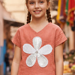 A young girl with braided hair wearing a terracotta-colored short-sleeve V-neck top with a large white sequined flower applique on the front, set against a blurred outdoor background.