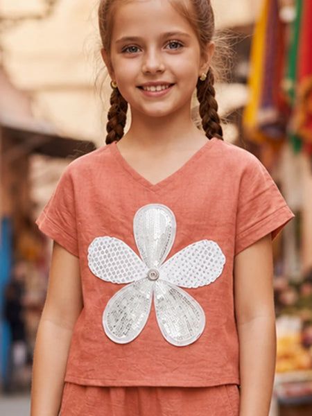 A young girl with braided hair wearing a terracotta-colored short-sleeve V-neck top with a large white sequined flower applique on the front, set against a blurred outdoor background.
