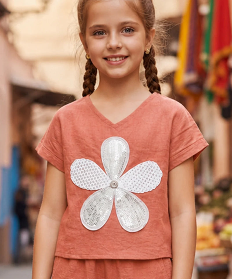 A young girl with braided hair wearing a terracotta-colored short-sleeve V-neck top with a large white sequined flower applique on the front, set against a blurred outdoor background.