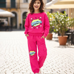 A young girl with curly brown hair stands on a cobblestone street wearing a bright pink two-piece tracksuit with a colorful comic-style graphic on the sweatshirt and pants. The background shows a blurred outdoor cafe setting with potted plants and buildings.