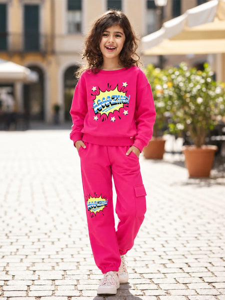 A young girl with curly brown hair stands on a cobblestone street wearing a bright pink two-piece tracksuit with a colorful comic-style graphic on the sweatshirt and pants. The background shows a blurred outdoor cafe setting with potted plants and buildings.