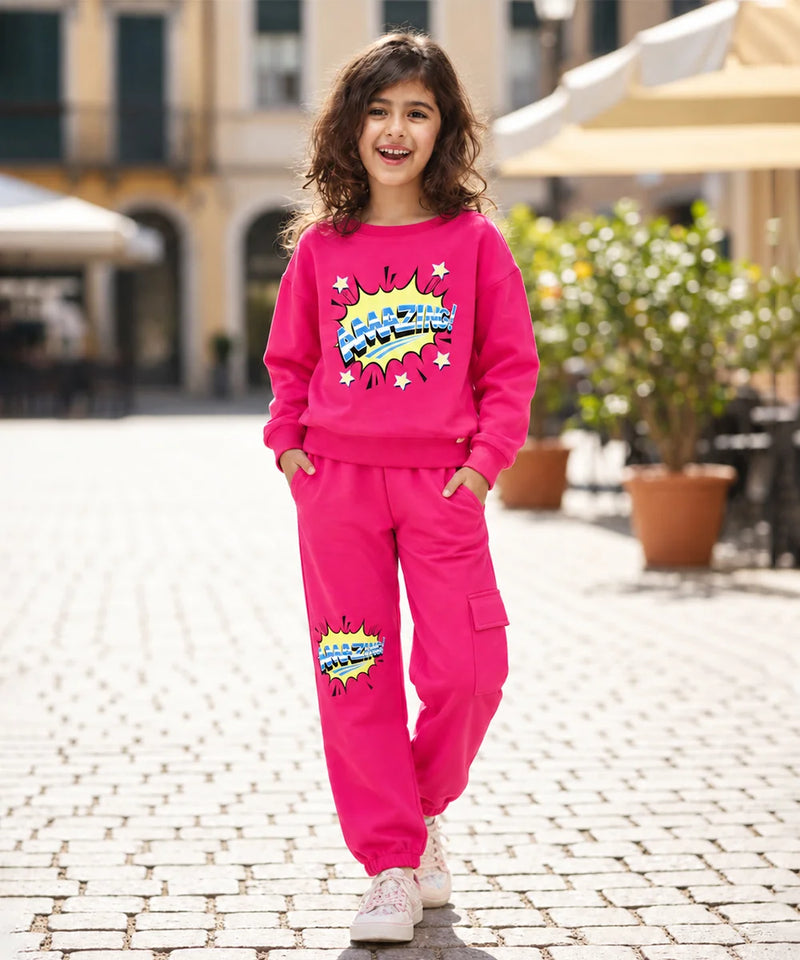 A young girl with curly brown hair stands on a cobblestone street wearing a bright pink two-piece tracksuit with a colorful comic-style graphic on the sweatshirt and pants. The background shows a blurred outdoor cafe setting with potted plants and buildings.