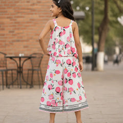 A young girl is shown from behind wearing a white sleeveless jumpsuit with a pink and green watercolor floral print. The jumpsuit features ruffled straps and a wide leg, with a black and white patterned hem that reads 'LEMONADE'.